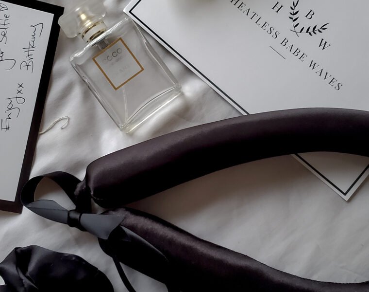 Black satin heatless curling ribbon, scrunchies, pearl hair clip and perfume arranged on a white bed